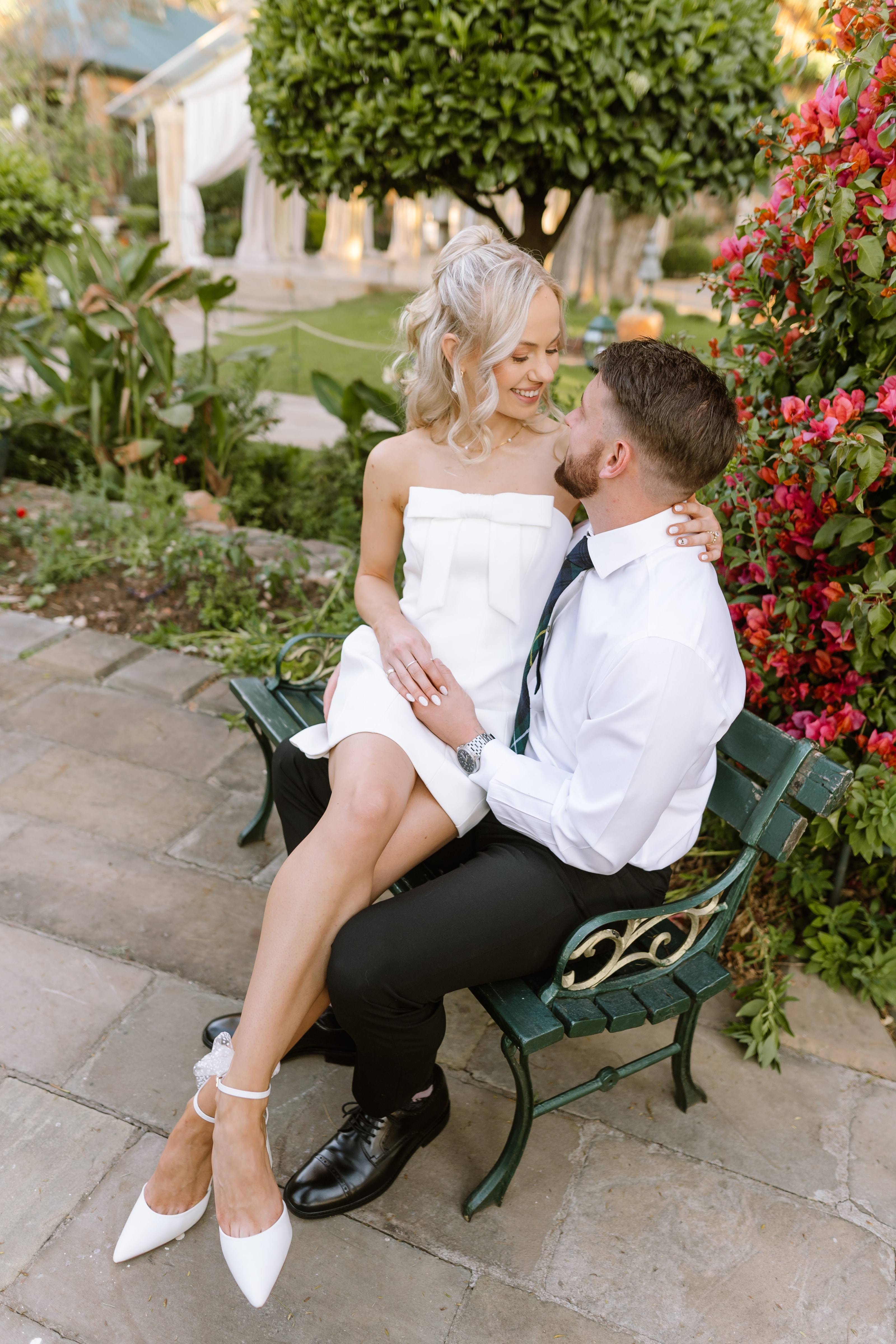 A bride in a white bow dress sits on her groom's lap outdoors