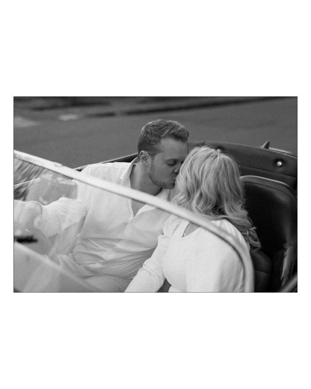 A couple in white attire shares a romantic kiss while sitting in a vintage convertible