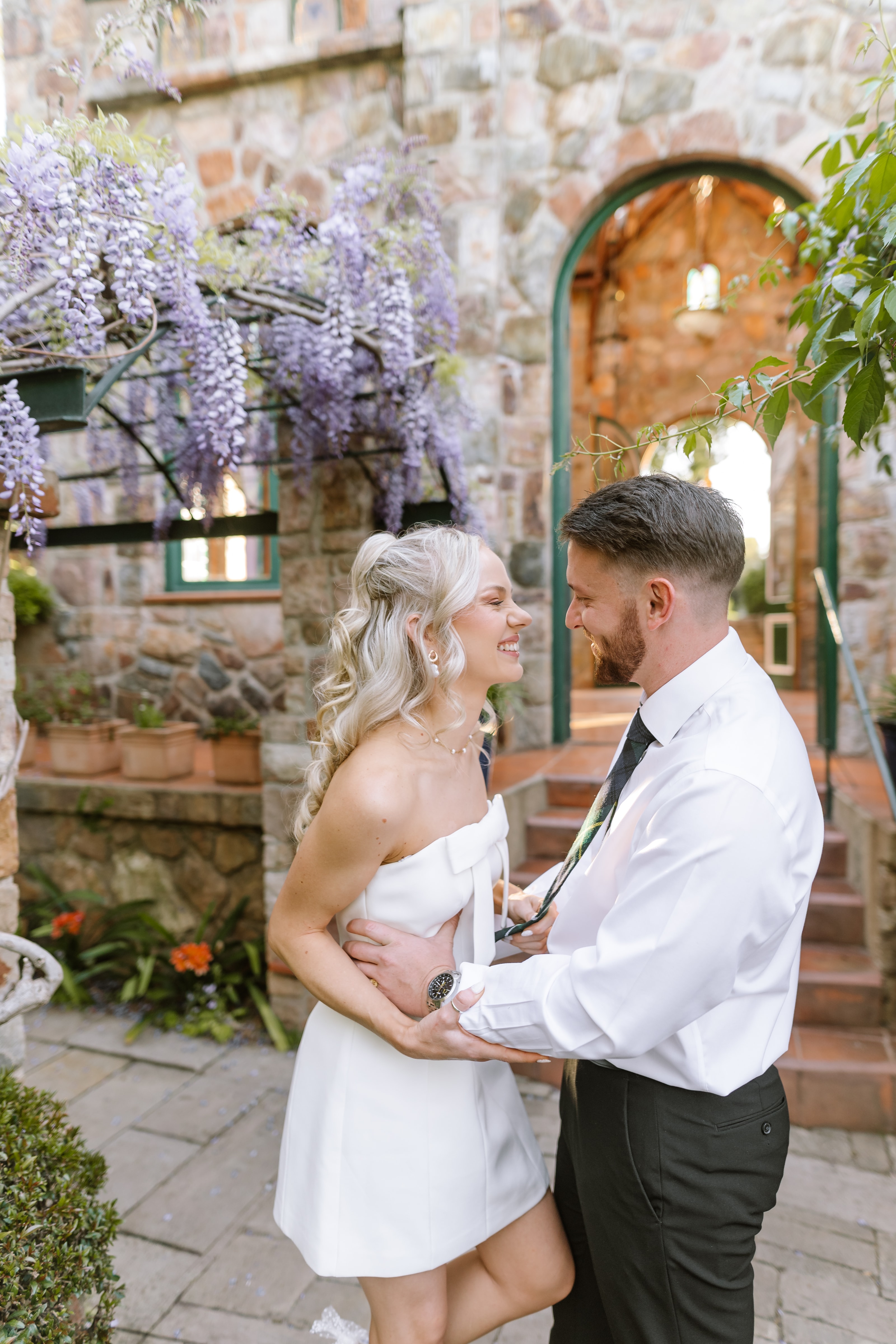 A joyful bride and groom share a romantic moment under blooming purple wisteria vines
