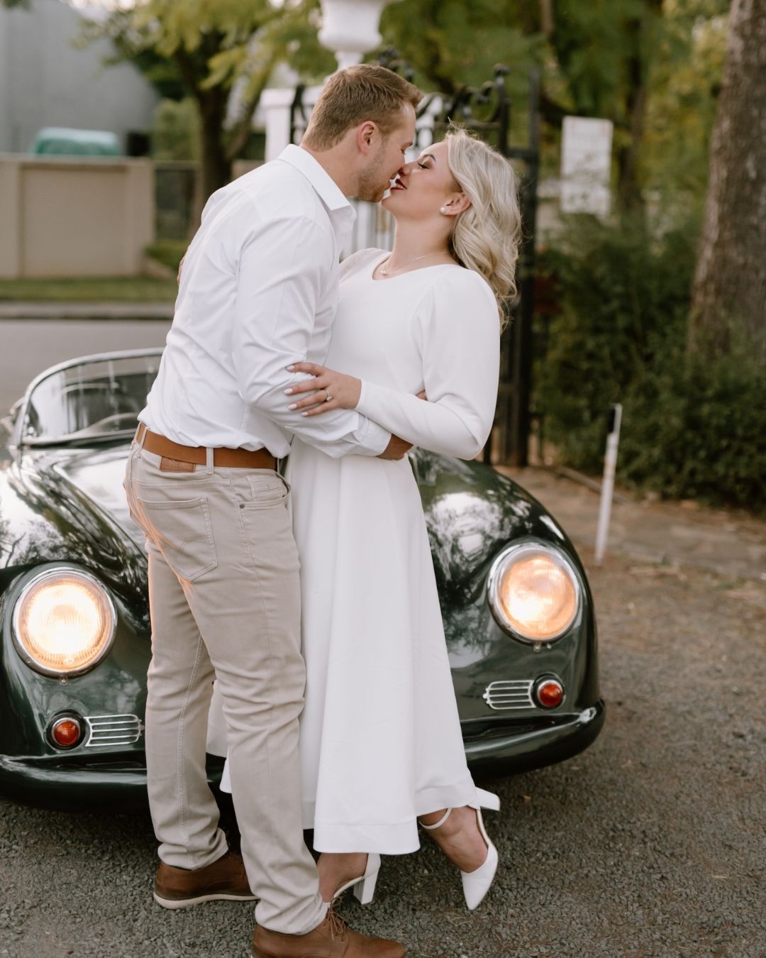 A couple in white wedding attire sharing a romantic kiss before a vintage green car