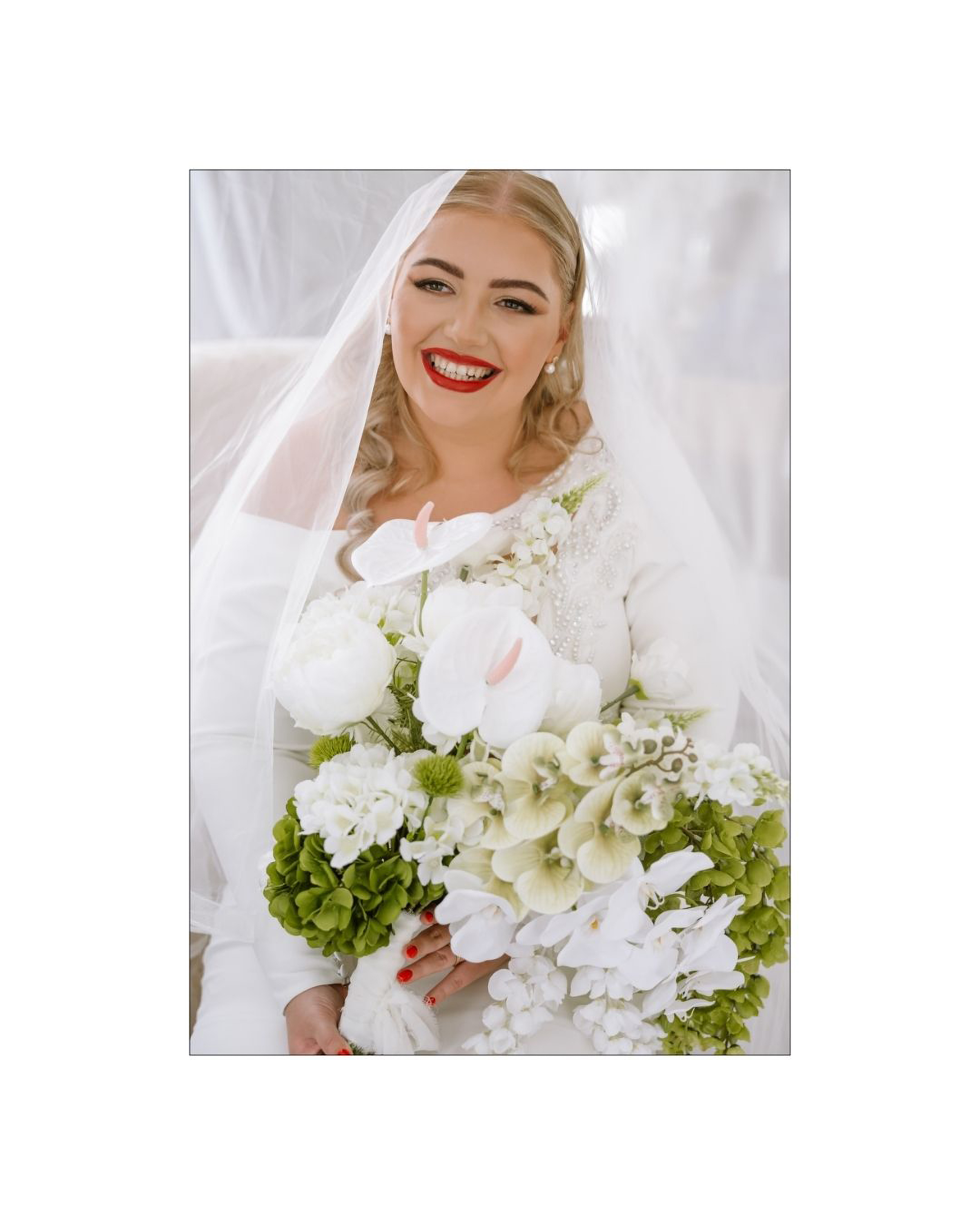 A smiling bride wearing a veil holds a large bouquet of white and green flowers