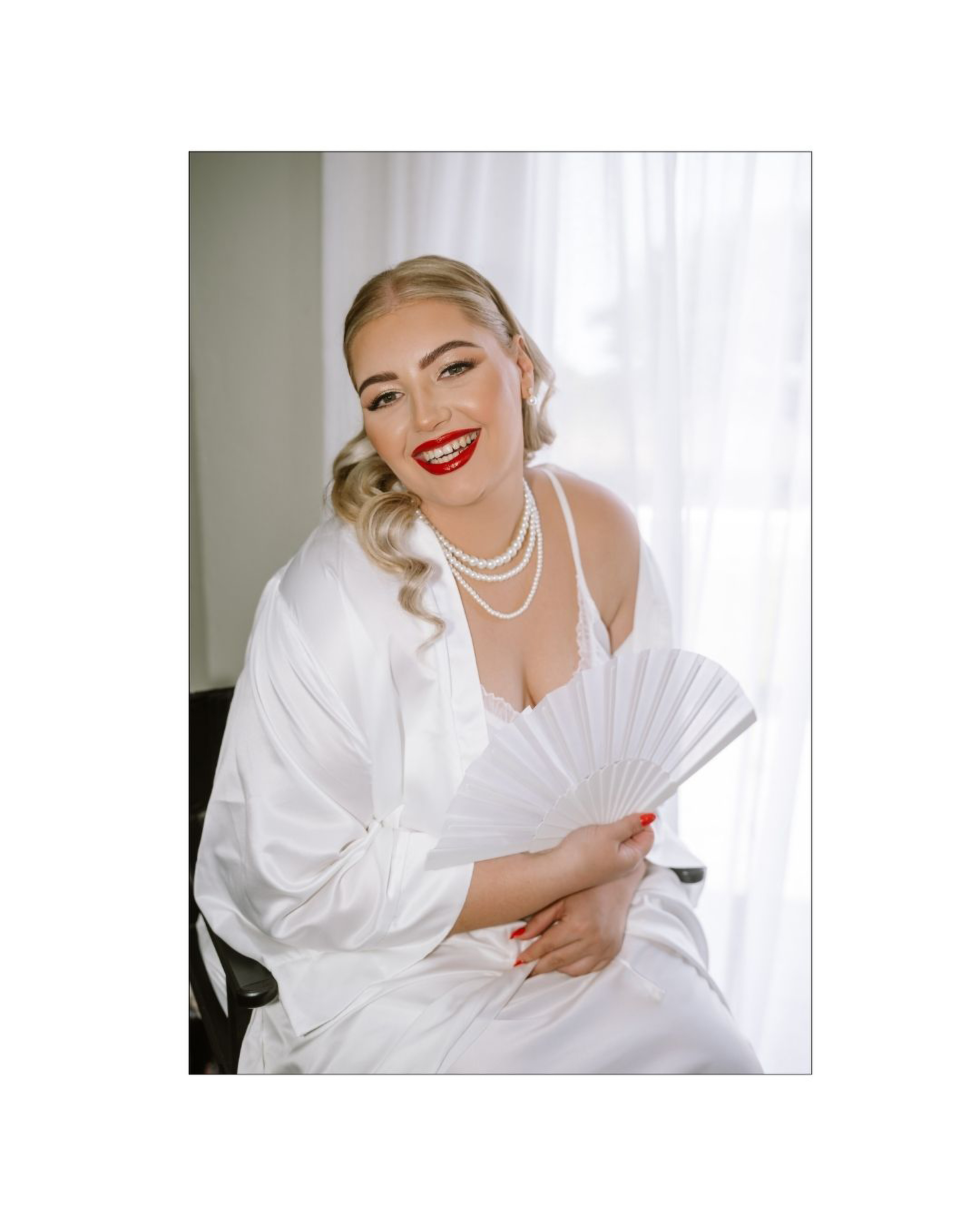 A smiling bride wearing a white robe and pearl necklace holds a hand fan