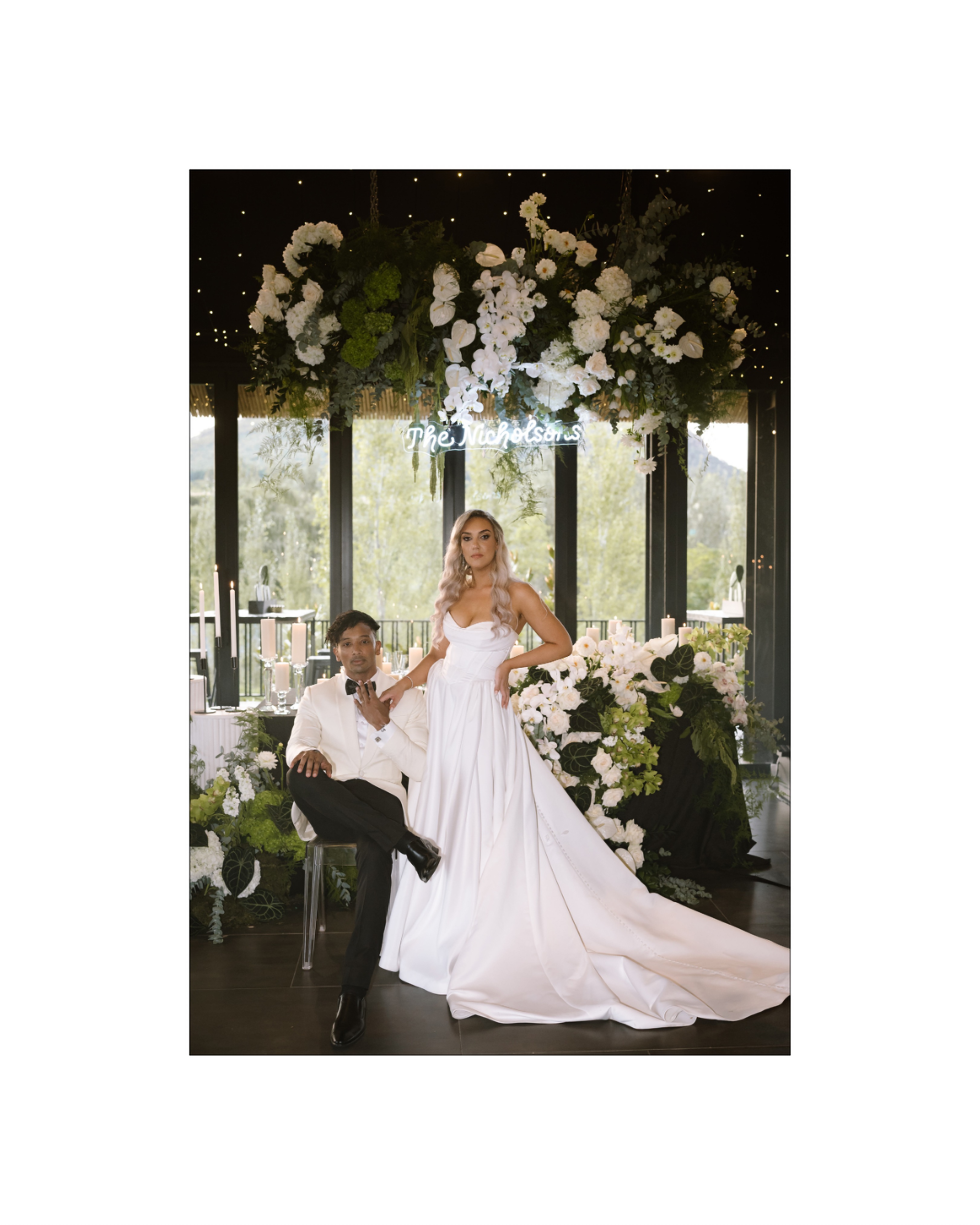 A stylish bride and groom pose together in front of a lush floral wedding installation