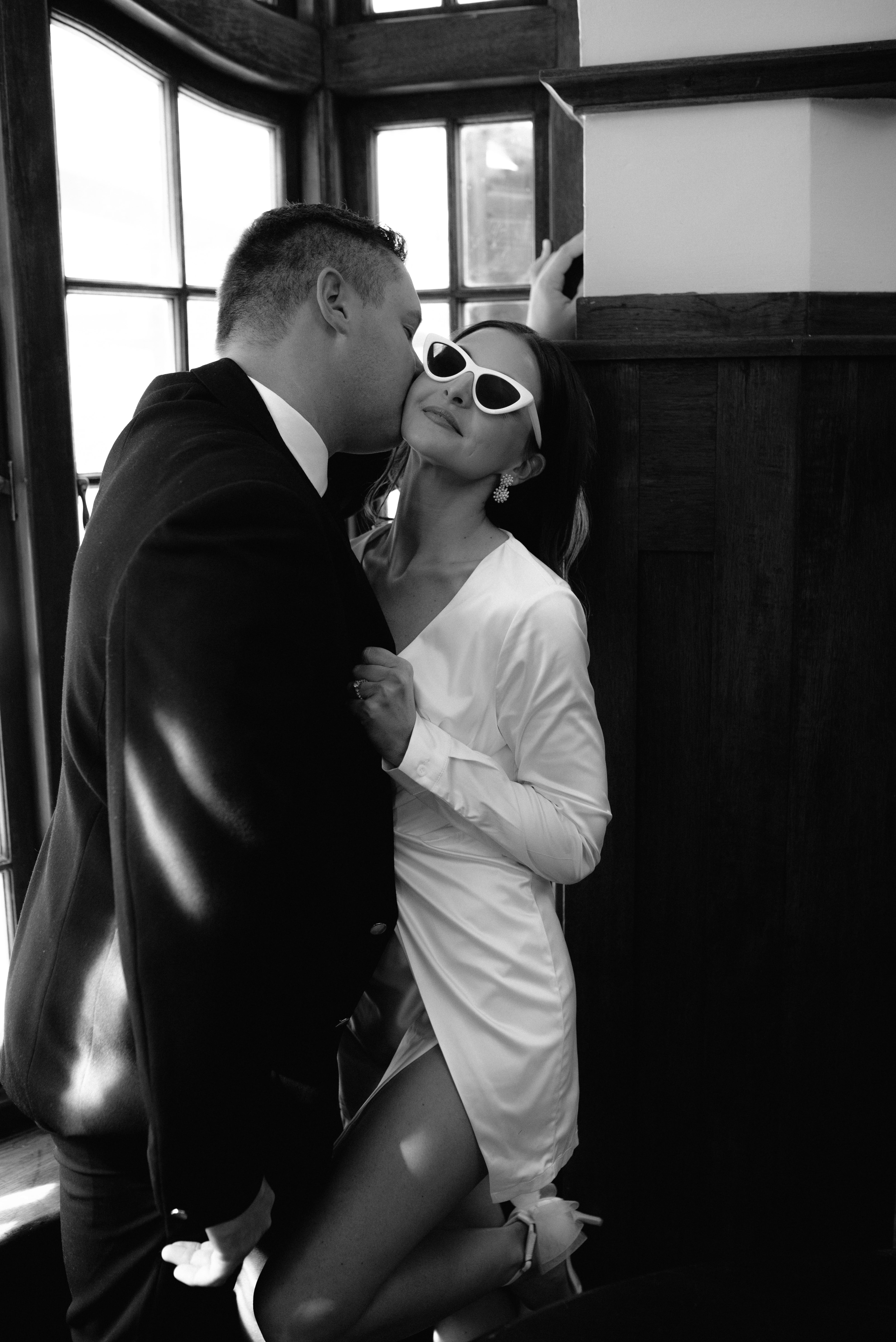 A stylish bride wearing white cat-eye sunglasses receives a kiss from her groom indoors