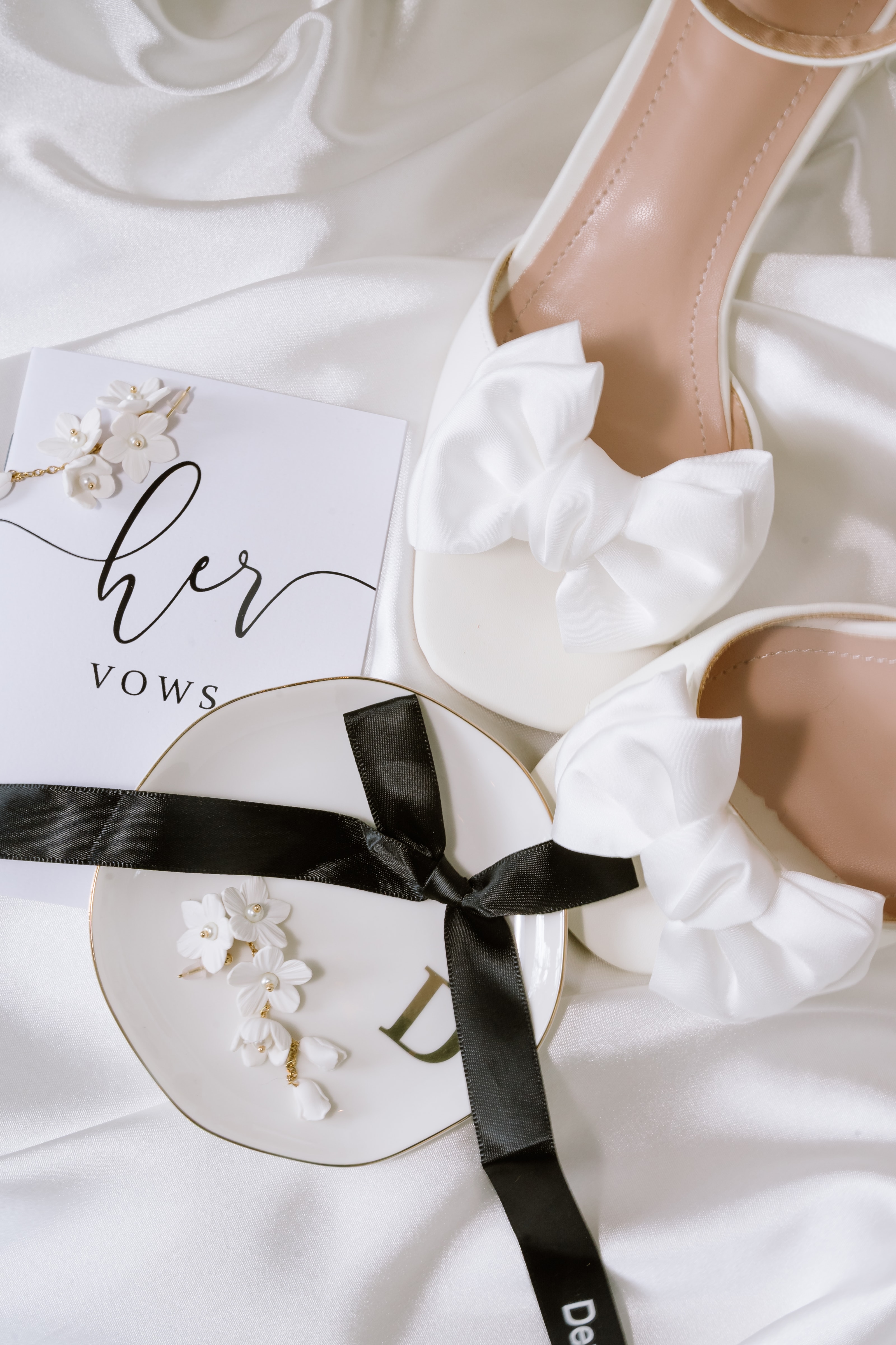 White satin bridal heels resting elegantly beside a vow book and delicate floral jewelry